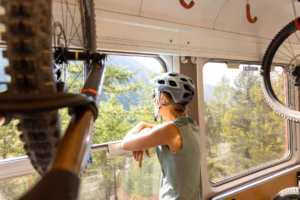 A woman wearing a helmet enjoys the view of a lush green landscape through a train window, with her mountain bike in view, during an outdoor adventure.
