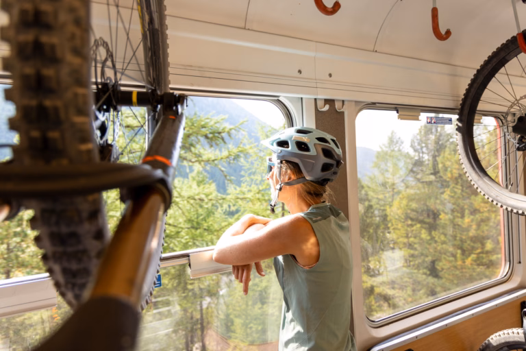 A woman wearing a helmet enjoys the view of a lush green landscape through a train window, with her mountain bike in view, during an outdoor adventure.